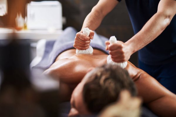 Close up of masseur hands massaging client back with herbal balls while he lying on massage table